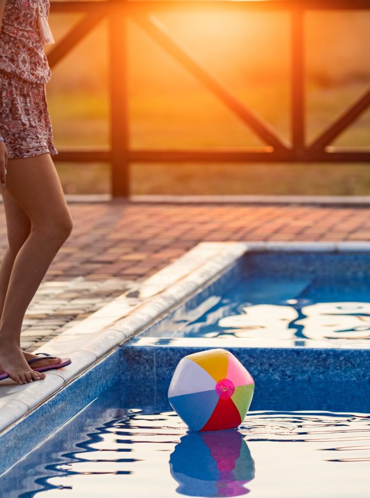 Fat tanned happy girl with dark hair braided in bun in bright summer suit is playing near pool with inflatable ball, against the background of golden summer evening sun