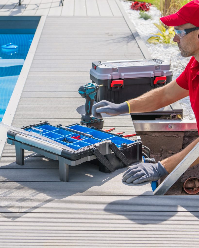 Outdoor Swimming Pool Heating and Filtering System Maintenance Performed by Caucasian Middle Aged Technician. Open Toolbox and Electric Drill Driver on the Floor in Front of Him.