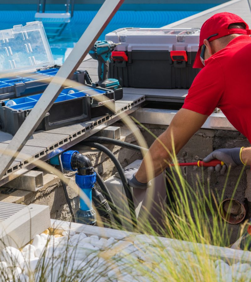 Closeup of Caucasian Professional Worker in Red Uniform Fixing Outdoor Swimming Pool Heating System Using Different Tools. Side View.