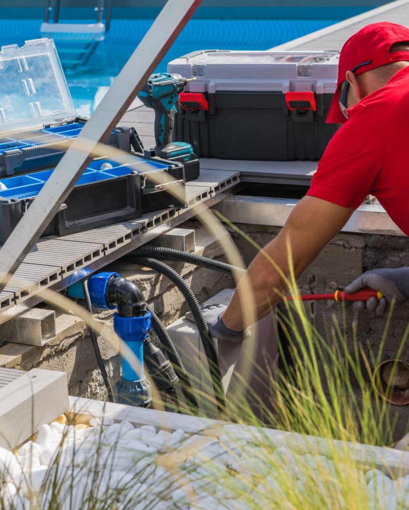 Closeup of Caucasian Professional Worker in Red Uniform Fixing Outdoor Swimming Pool Heating System Using Different Tools. Side View.